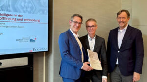 Drei Männer in Anzügen stehen nebeneinander, einer hält eine Urkunde, im Hintergrund ein Bildschirm auf dem eine Präsentation abgespielt wird