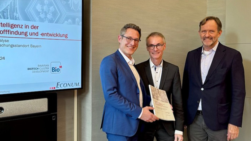 Drei Männer in Anzügen stehen nebeneinander, einer hält eine Urkunde, im Hintergrund ein Bildschirm auf dem eine Präsentation abgespielt wird