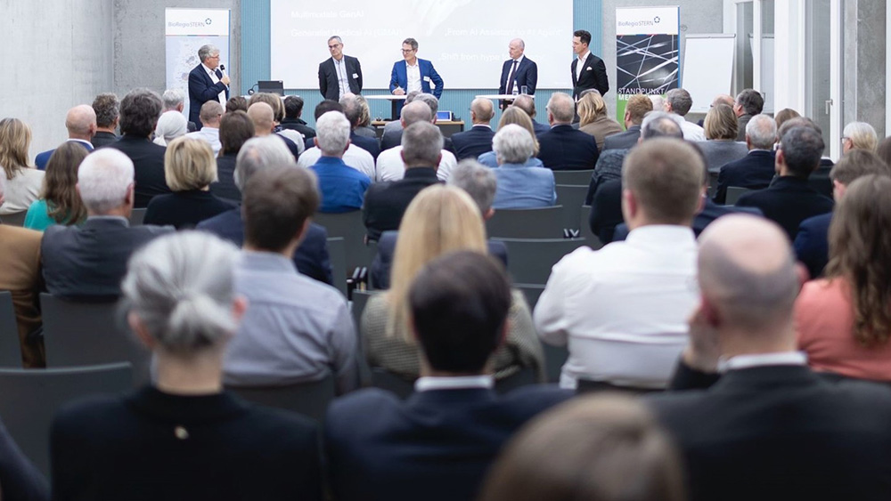 Publikum sitzt in einem Saal und hört einem Podium auf dem fünf Personen sprechen, zu