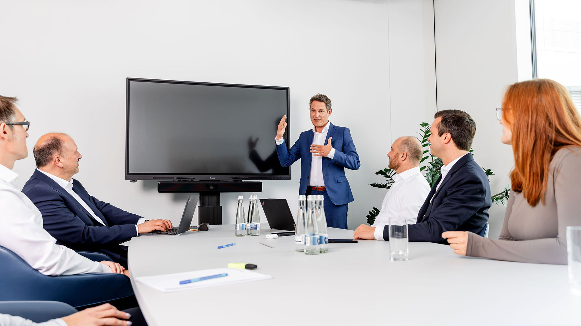 Geschäftsmann in blauem Anzug steht vor großem Bildschirm und präsentiert vor fünf sitzenden Personen in einem modernen Konferenzraum