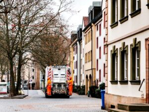 Ein Müllwagen mit zwei orange gekleideten Arbeitern an der Rückseite fährt eine gepflasterte Straße mit mehrstöckigen Wohnhäusern entlang