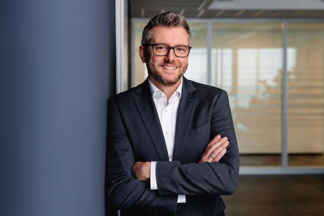 Lockeres Portrait Gregor Böll, angelehnt an blauer Wand